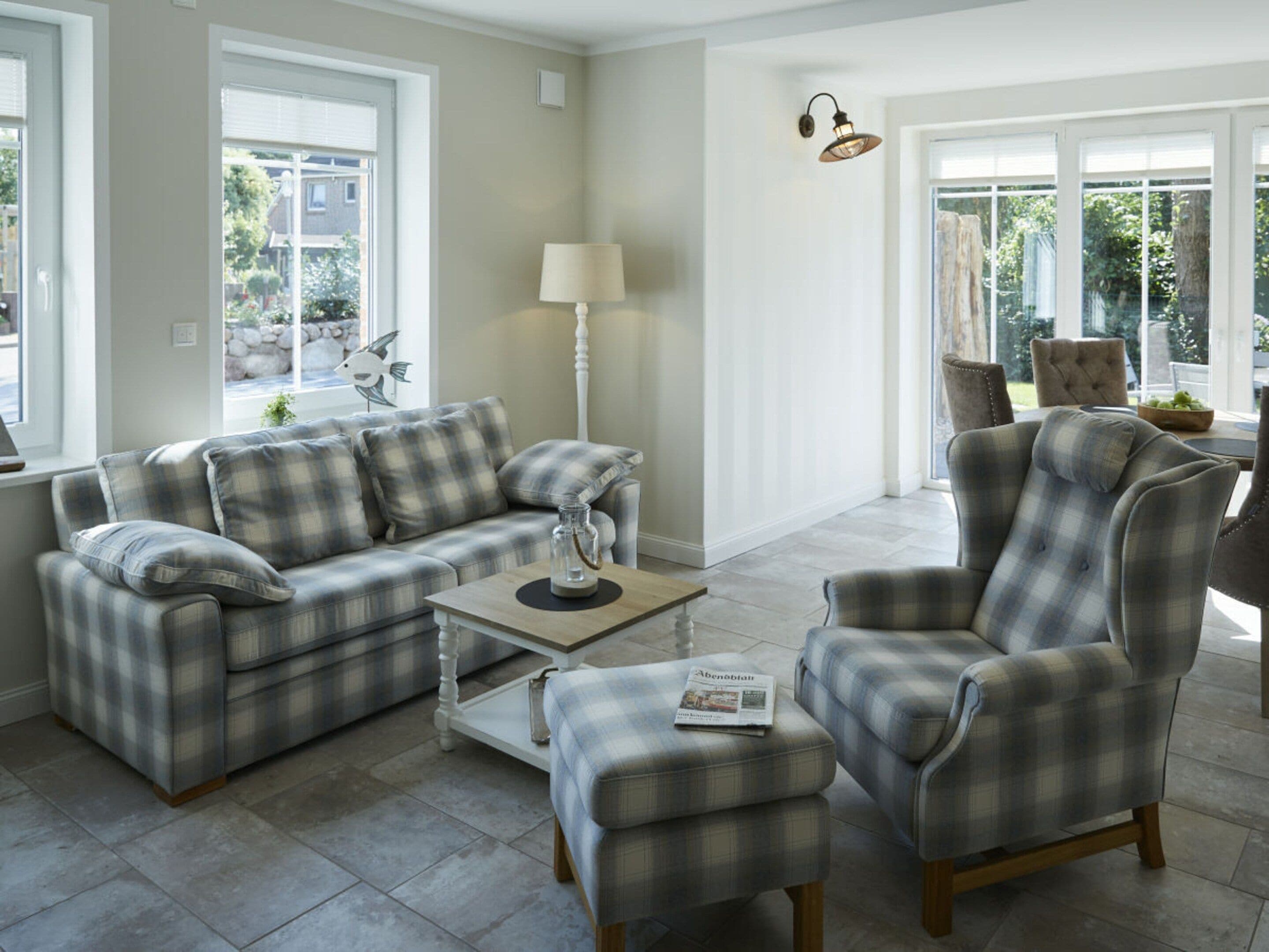 A living room with a plaid sofa, armchair, and ottoman, a lamp, and a table with a newspaper.
