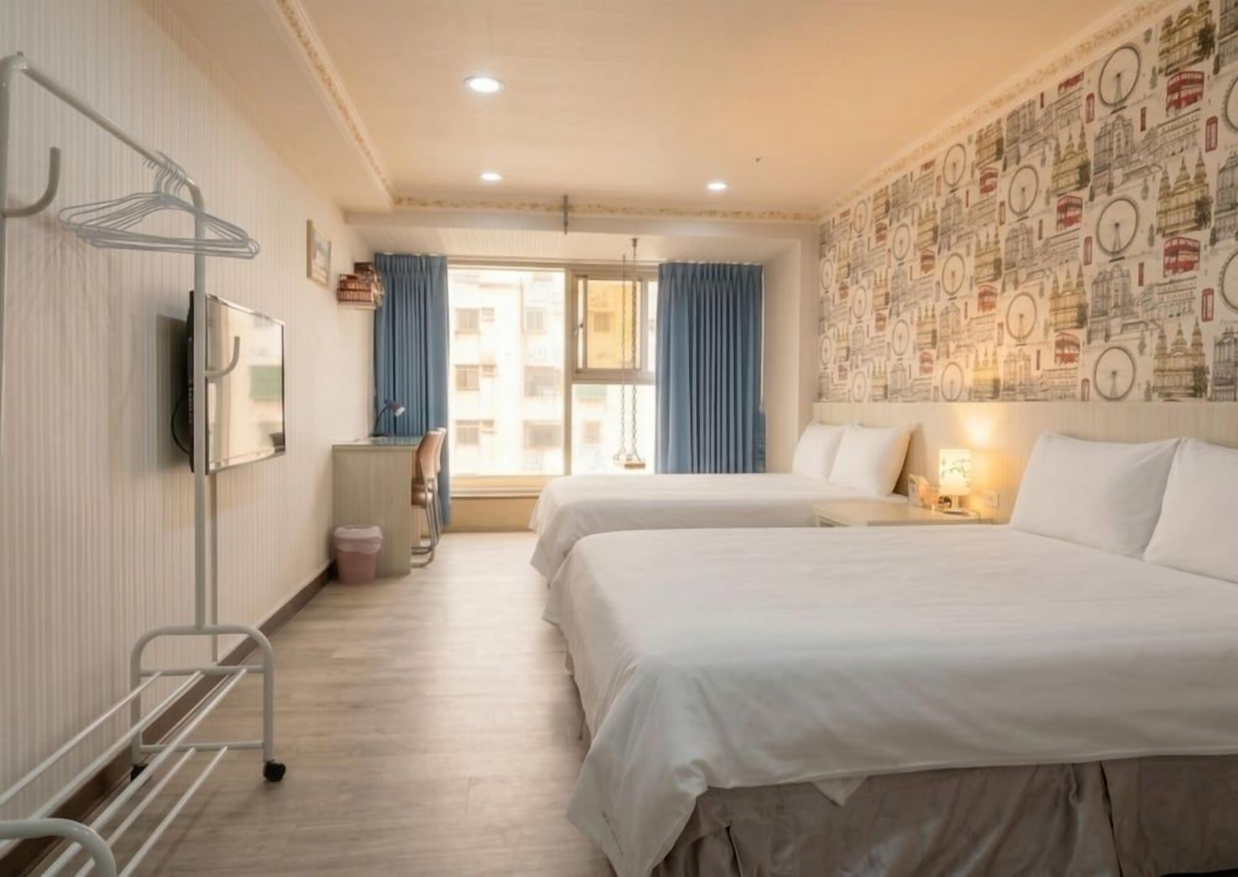 Ein Hotelzimmer mit zwei Betten, einem Fernseher, einem Fenster mit Vorhängen und einer Wand mit Stadtansicht-Muster.