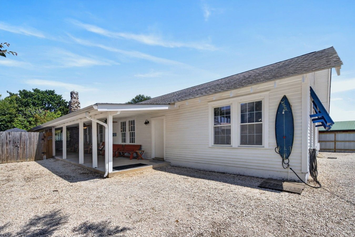 Une maison de plain-pied avec un patio couvert, une planche de surf appuyée contre le mur et une clôture en bois en arrière-plan.