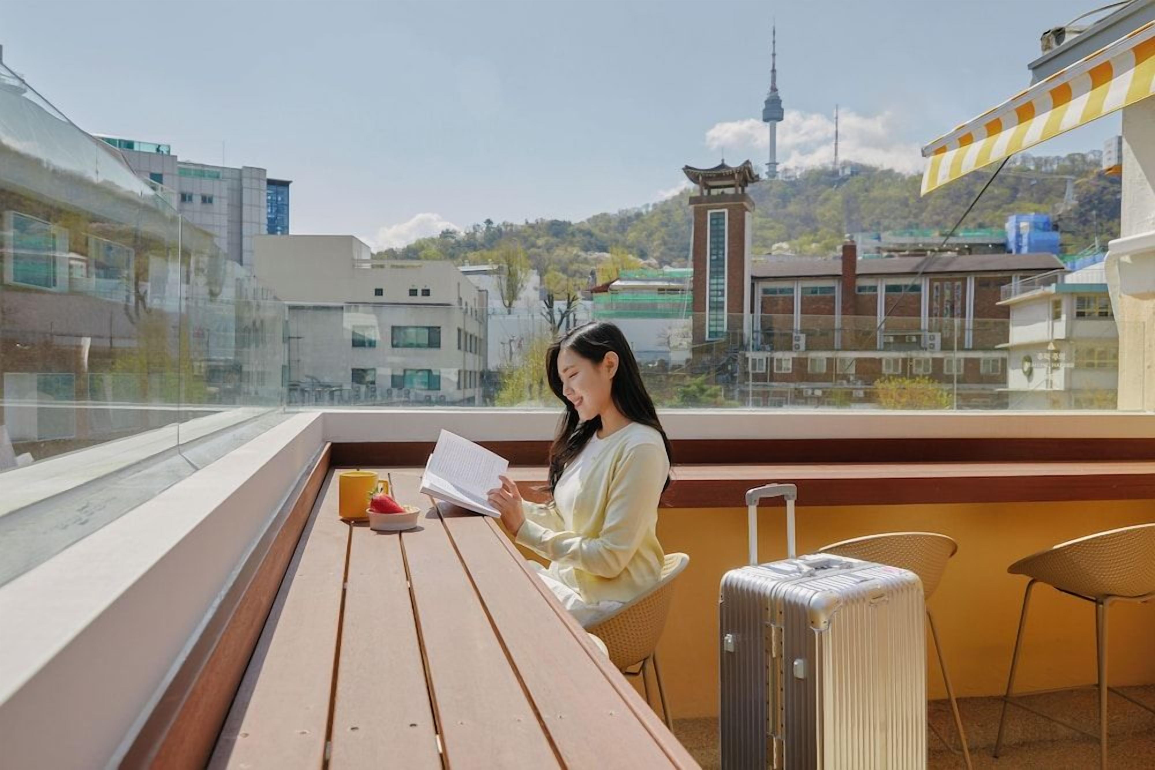 A person sitting at a wooden table with a book, a suitcase, and a cup on the balcony overlooking a cityscape with buildings and a tower.