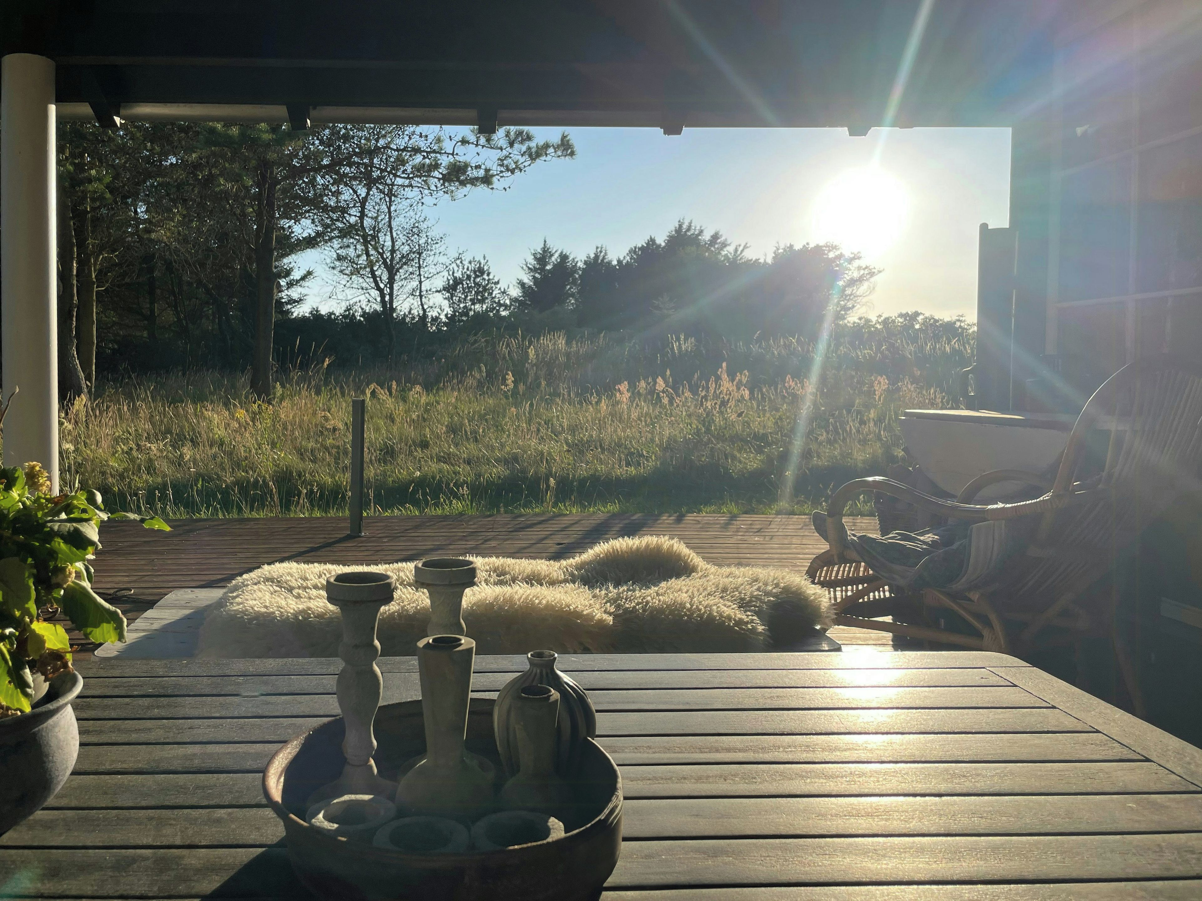 Eine Holzterrasse mit einer rustikalen Sitzgruppe im Freien, einer Schale mit dekorativen Vasen und Blick auf ein Wiesenfeld und Bäume.