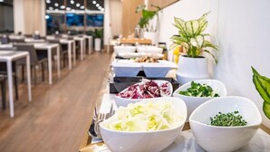 Een buffetopstelling met diverse gerechten in schalen, een eetruimte met tafels en stoelen, en potplanten.