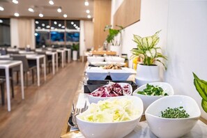 En buffé med olika rätter upplagda i skålar, ett matområde med bord och stolar samt krukväxter.