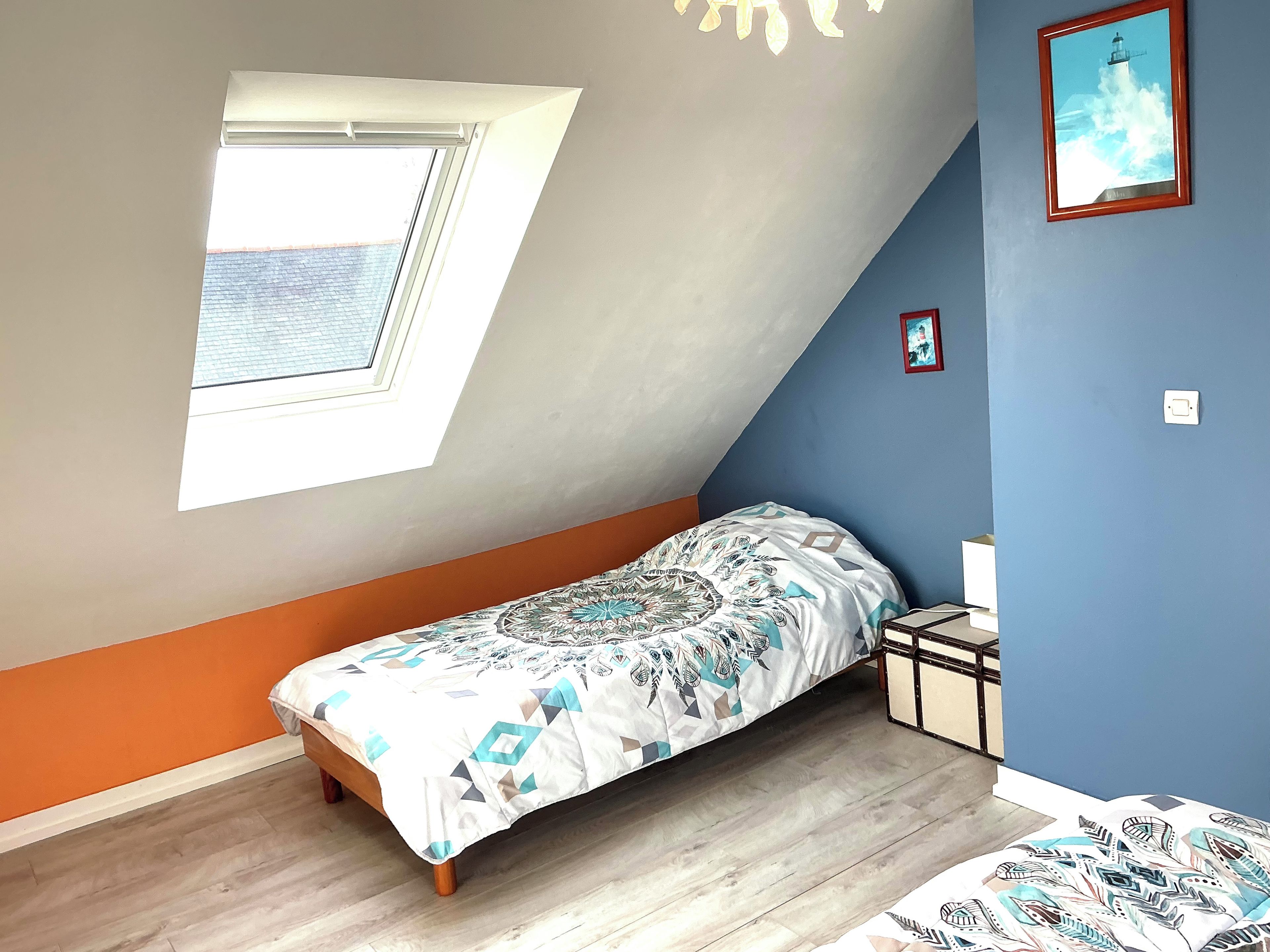 A small, single-bed room with a sloped ceiling, a skylight, a patterned bedspread, and a framed picture on the wall.