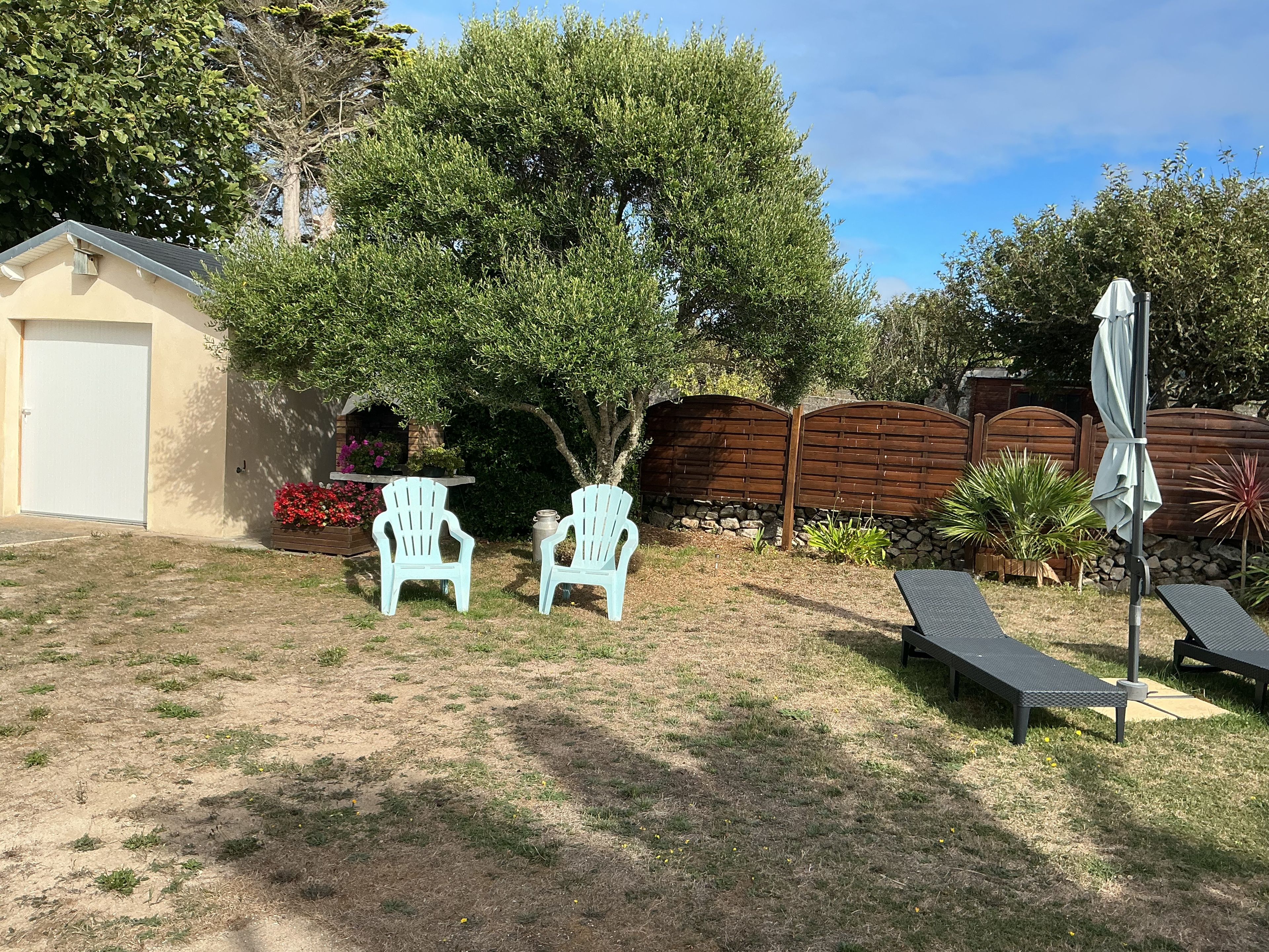 Ein Garten mit Garage, zwei Adirondack-Stühlen, einem kleinen Tisch, einem Metall-Sonnenschirm und einem Holzzaun.