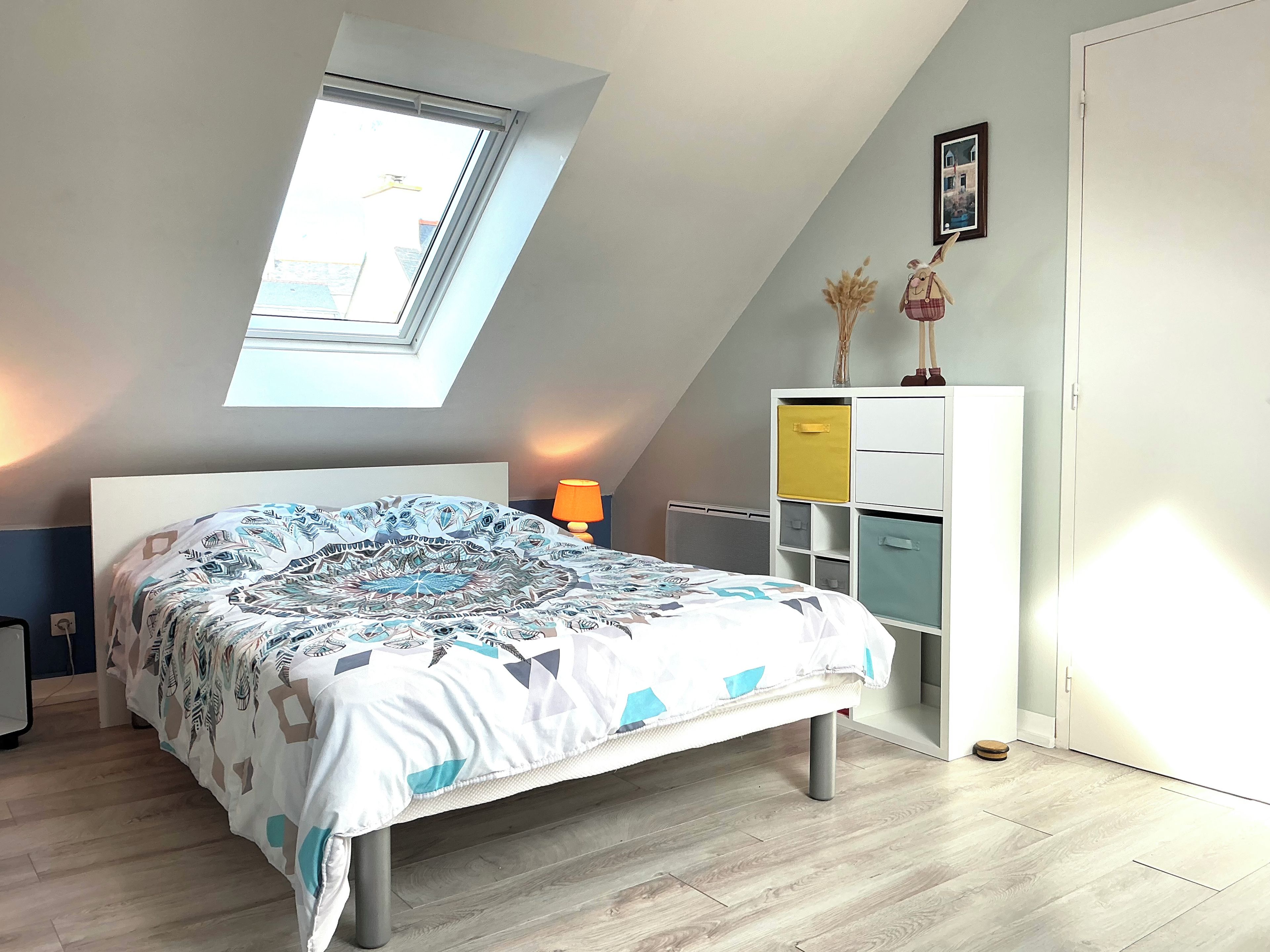 Un dormitorio con techo inclinado, tragaluces, cama con ropa de cama estampada, lámpara de noche, cómoda con varios cajones y radiador.