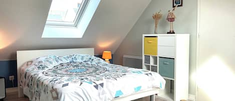 Ein Schlafzimmer mit schrägem Dach, einem Oberlicht, einem Bett mit gemusterten Betttextilien, einer Nachttischlampe, einer Kommode mit mehreren Schubladen und einem Heizkörper.