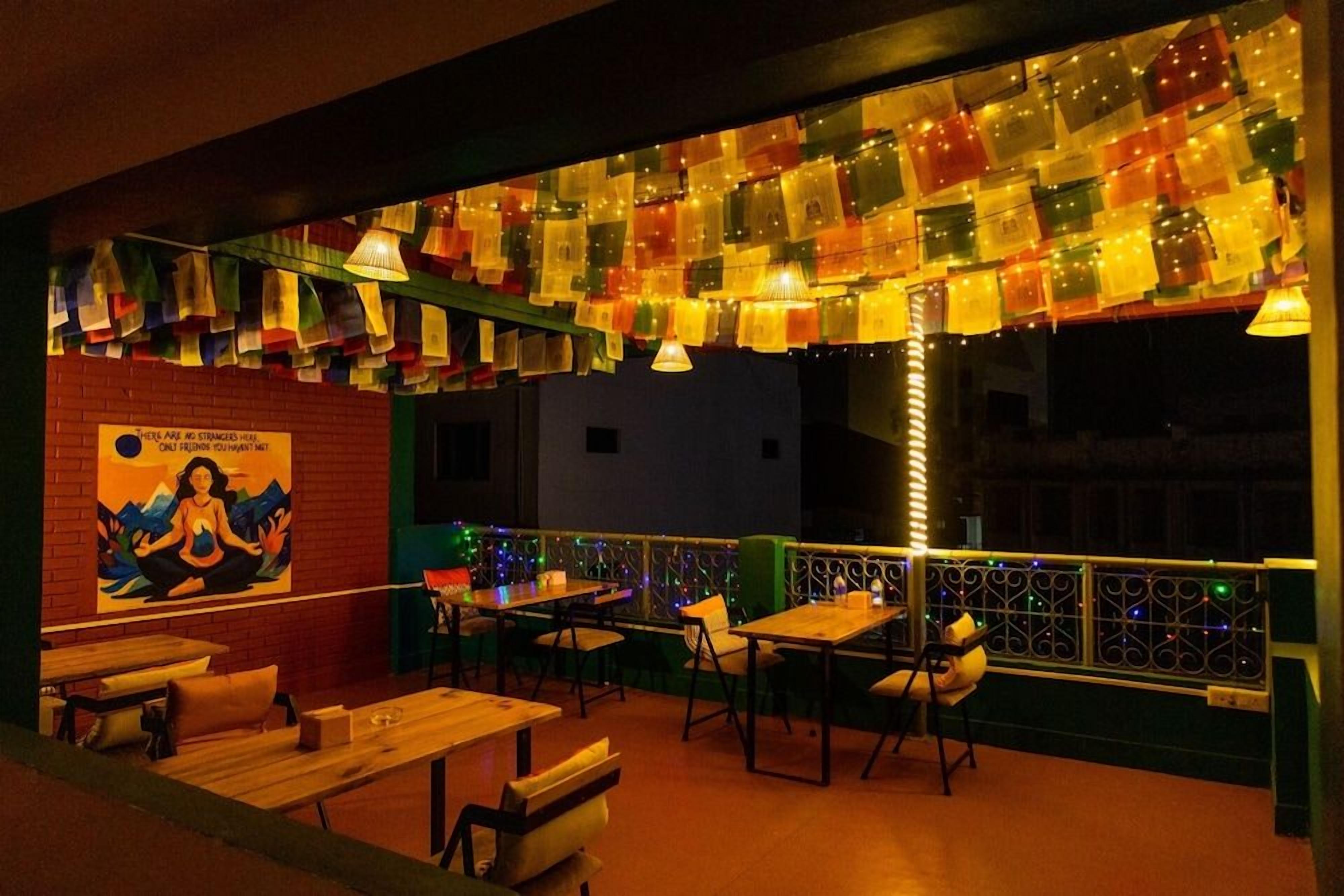 An outdoor seating area with tables and chairs, a mural on the wall, and colorful lights strung above.