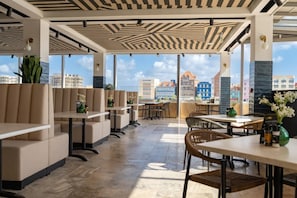 A modern outdoor seating area with tables and chairs, large windows offering a view of buildings, and a patterned ceiling. - Harbor Hotel and Casino Curacao (Willemstad)