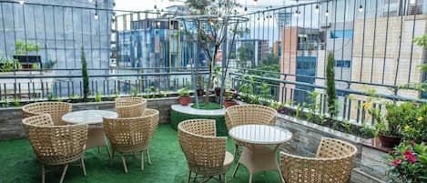 Una terrazza esterna con mobili in vimini e un piccolo albero, circondata da un recinto metallico e con edifici urbani sullo sfondo.
