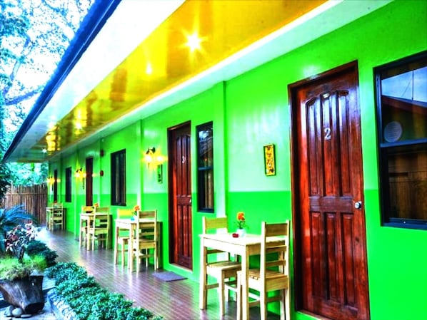 Un edificio con fachada verde, puertas de madera y un porche cubierto con sillas y mesas.