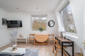 A modern, well-lit room with a flat-screen TV mounted on the wall, a dining table with chairs, a small side table with a lamp, and a large window providing a view of trees. - Top-Floor 1-Bed Apt. in South Kensington (London)