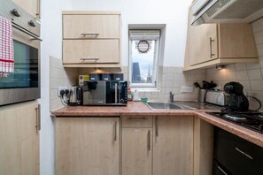 A compact kitchen with a microwave, toaster, and coffee maker on the countertop, a window with a view of a cityscape, and light-colored cabinets. - Top-Floor 1-Bed Apt. in South Kensington (London)