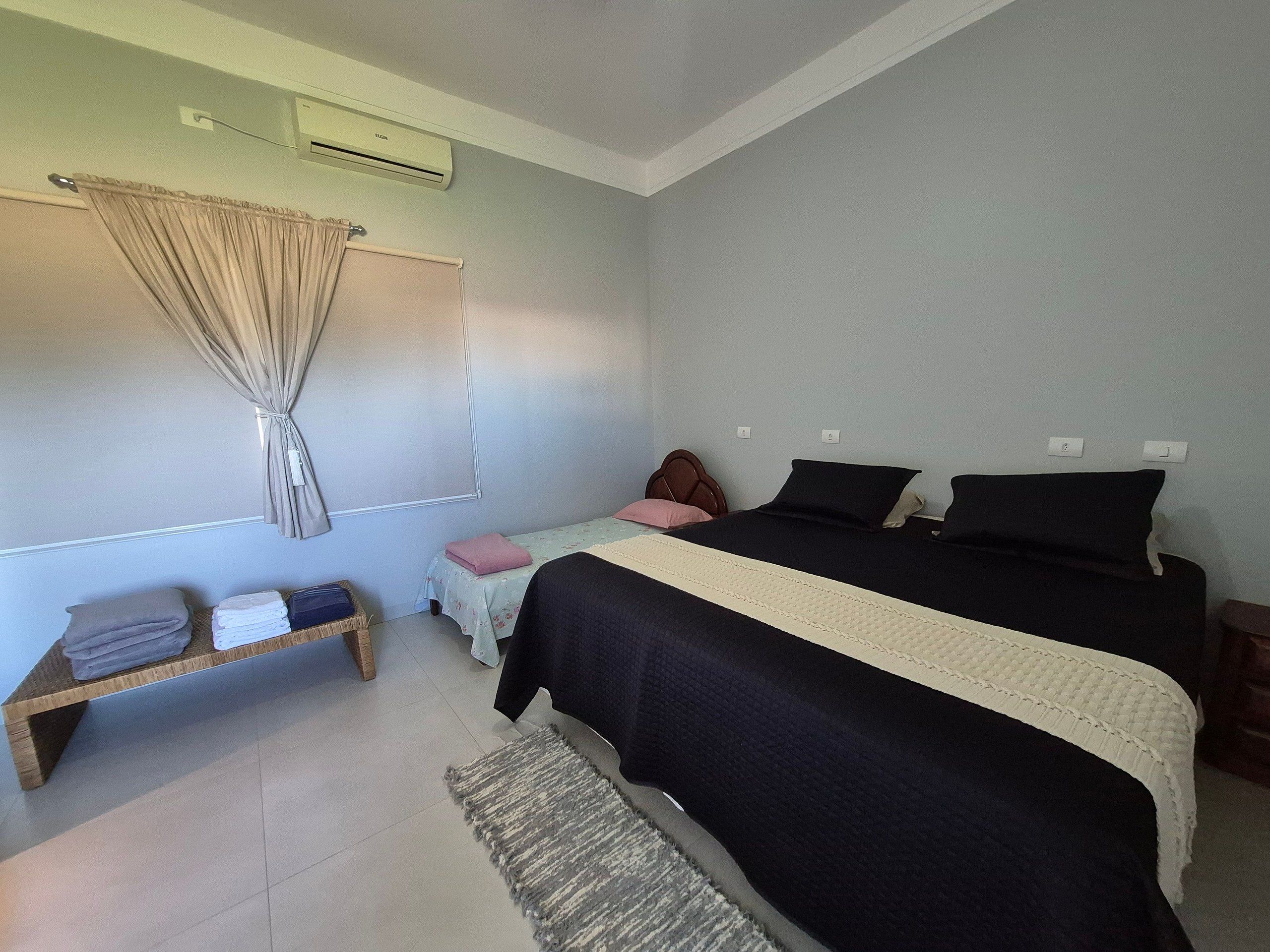 Un dormitorio con cama, una mesita de noche, una pequeña mesa con toallas dobladas, aire acondicionado de pared y una ventana con cortinas.