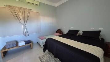 Un dormitorio con cama, una mesita de noche, una pequeña mesa con toallas dobladas, aire acondicionado de pared y una ventana con cortinas.