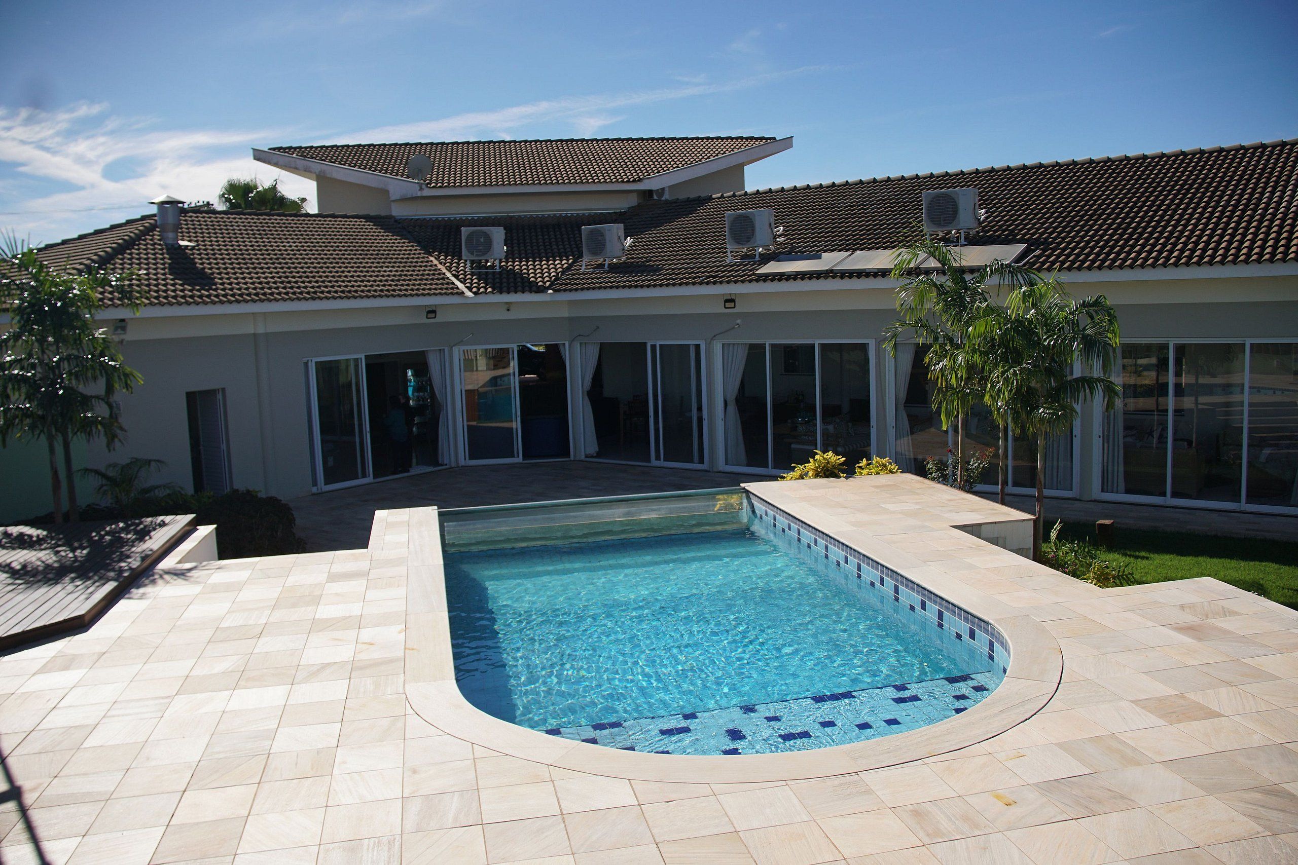 Uma piscina com deck ladrilhado, uma casa com telhado telhado e janelas grandes.