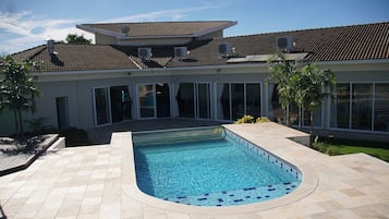 Una piscina con bordeado de baldosas, una casa con techo de tejas y ventanas amplias.