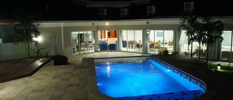 A swimming pool with a tiled deck, a covered patio area, and a view into the building.