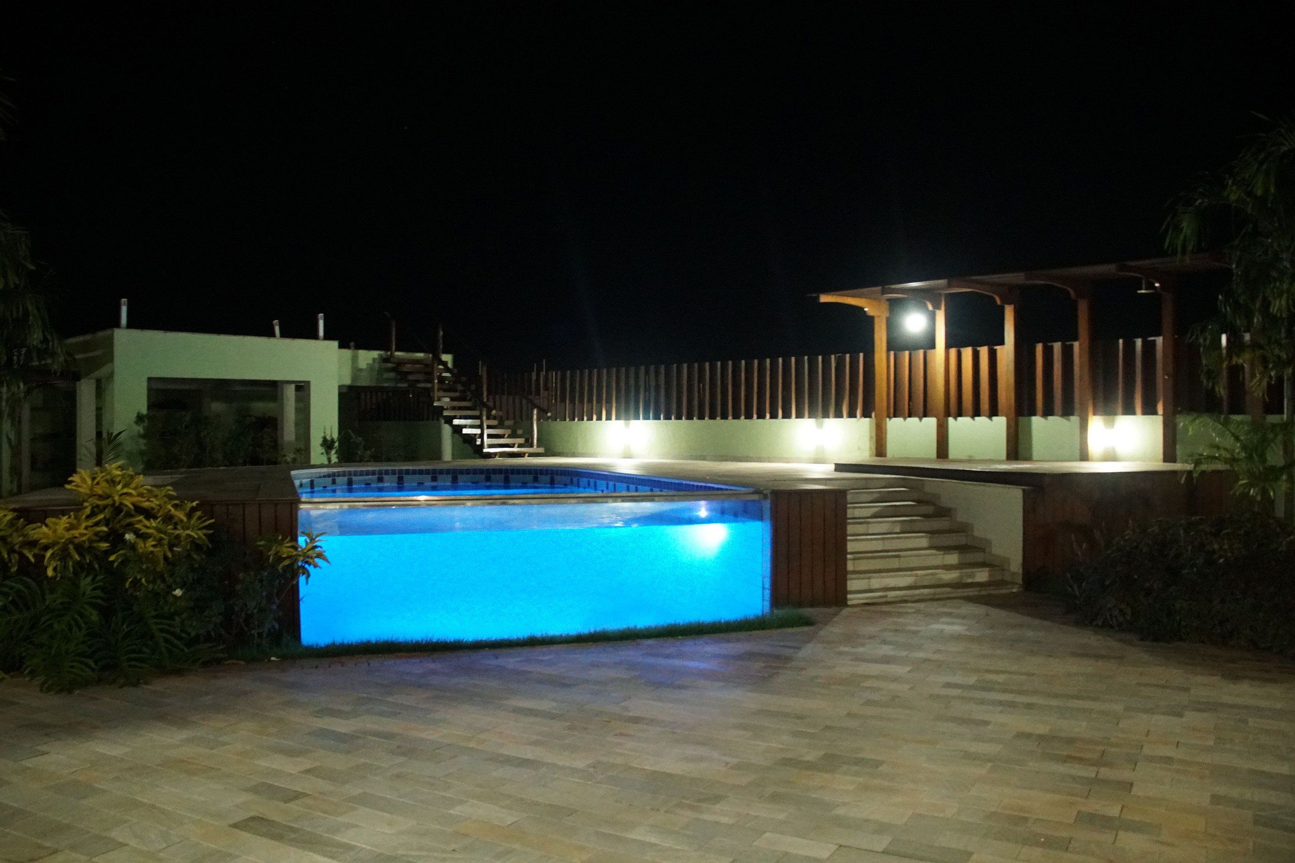 Piscina com borda iluminada, pérgola de madeira e escadaria para um segundo andar.