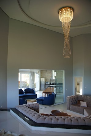A spacious living area with a high ceiling, a chandelier, and a view into a kitchen through a glass door.