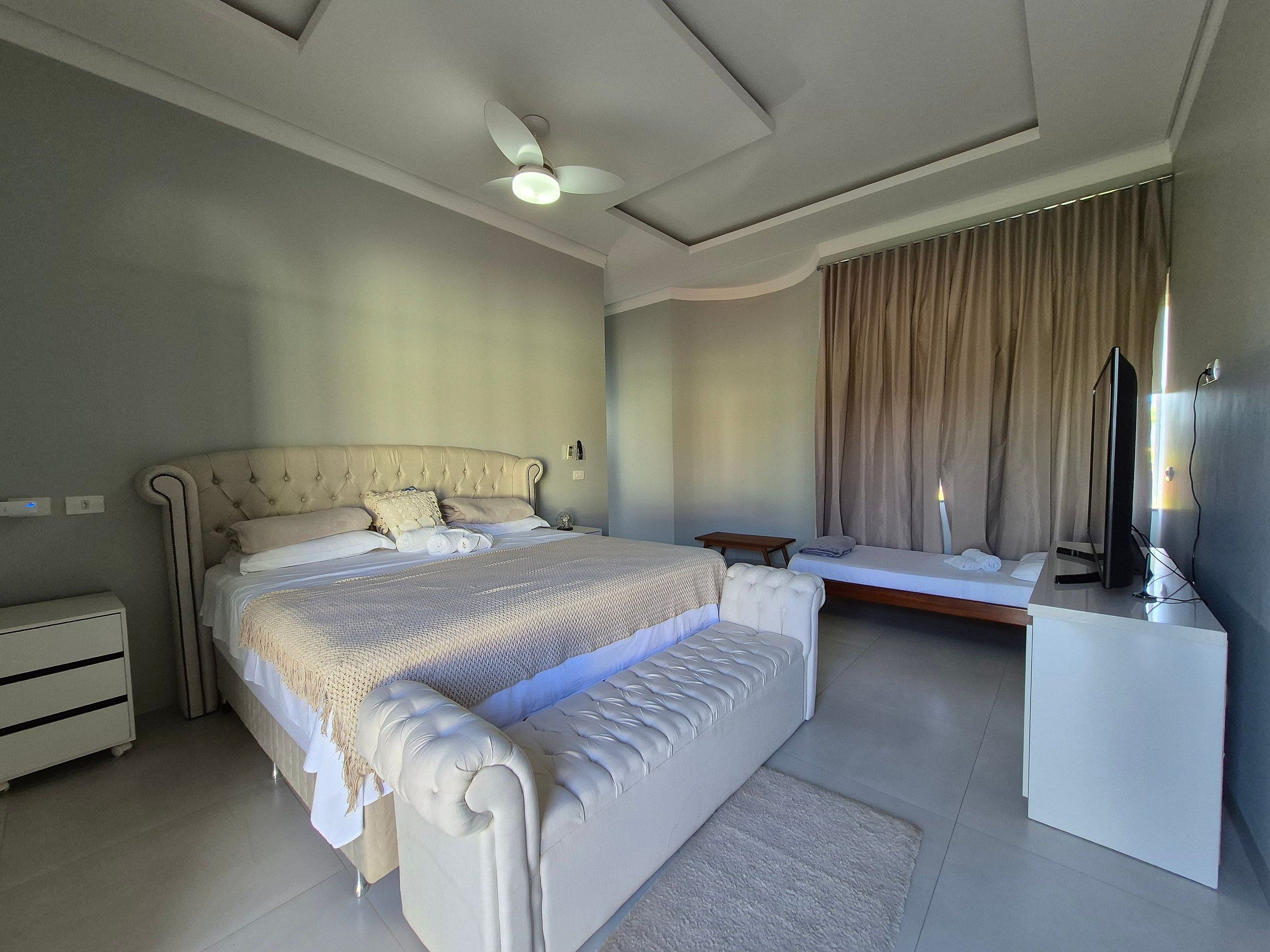 A bedroom with a large bed, a bedside table, a desk with a computer, a ceiling fan, and a window with curtains.