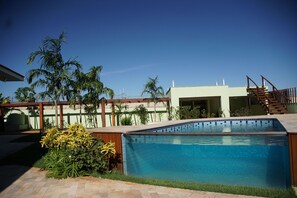 Una piscina con borde de azulejos, rodeada de vegetación y un edificio con escaleras.