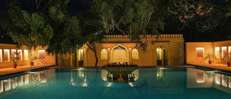 A nighttime pool area with a fountain, trees, and traditional architecture.
