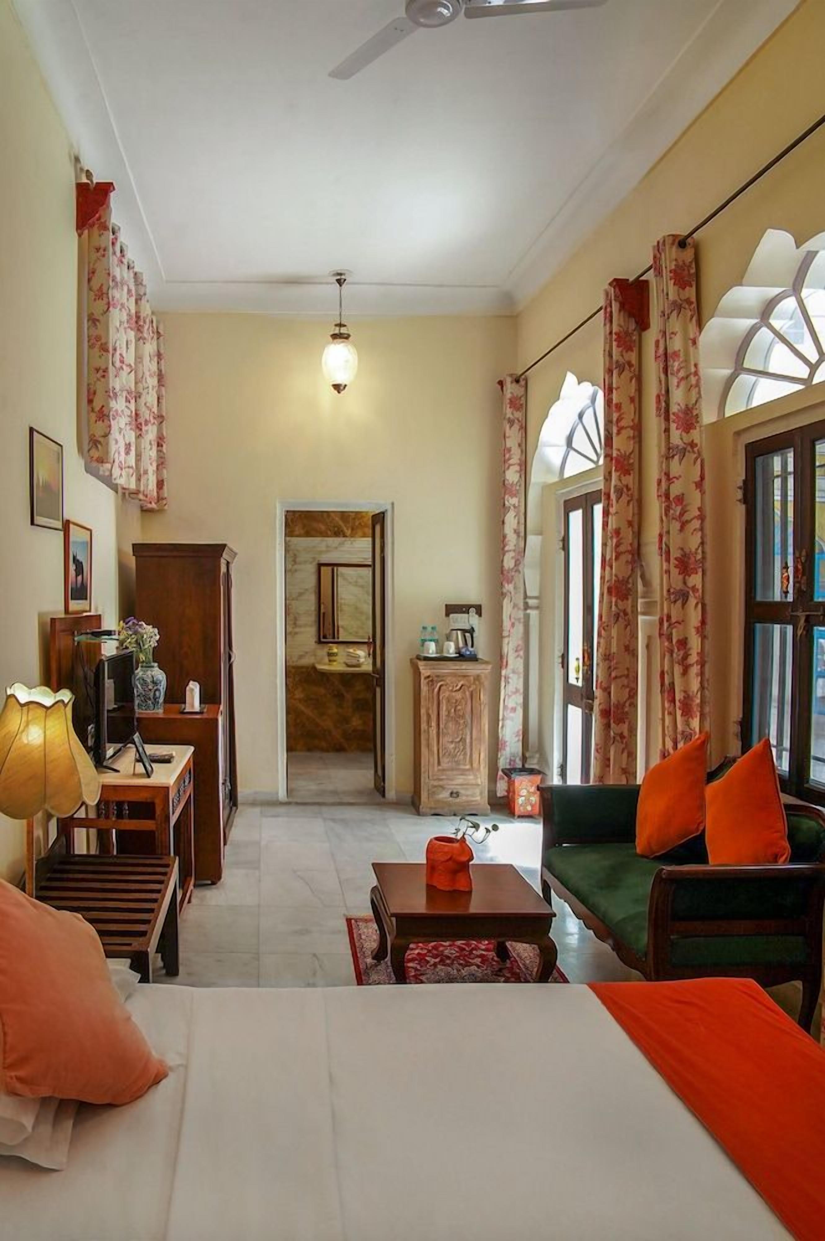 A hotel room with a bed, a sofa, a coffee table, a desk with a lamp, and a television. There is a window with curtains and a door leading to a bathroom.