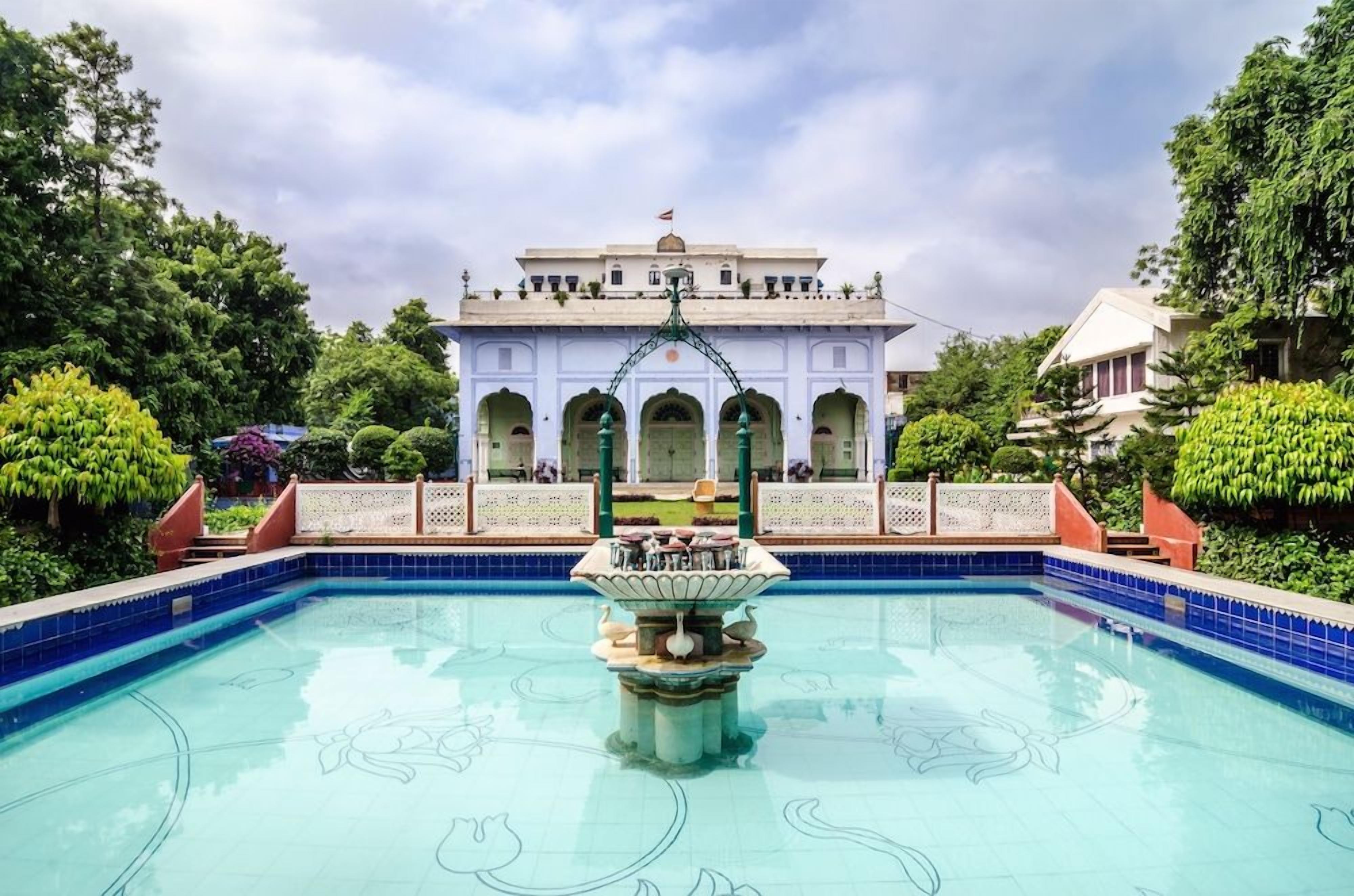A swimming pool with a fountain in the center, surrounded by a garden with neatly trimmed bushes and a building with arches in the background.