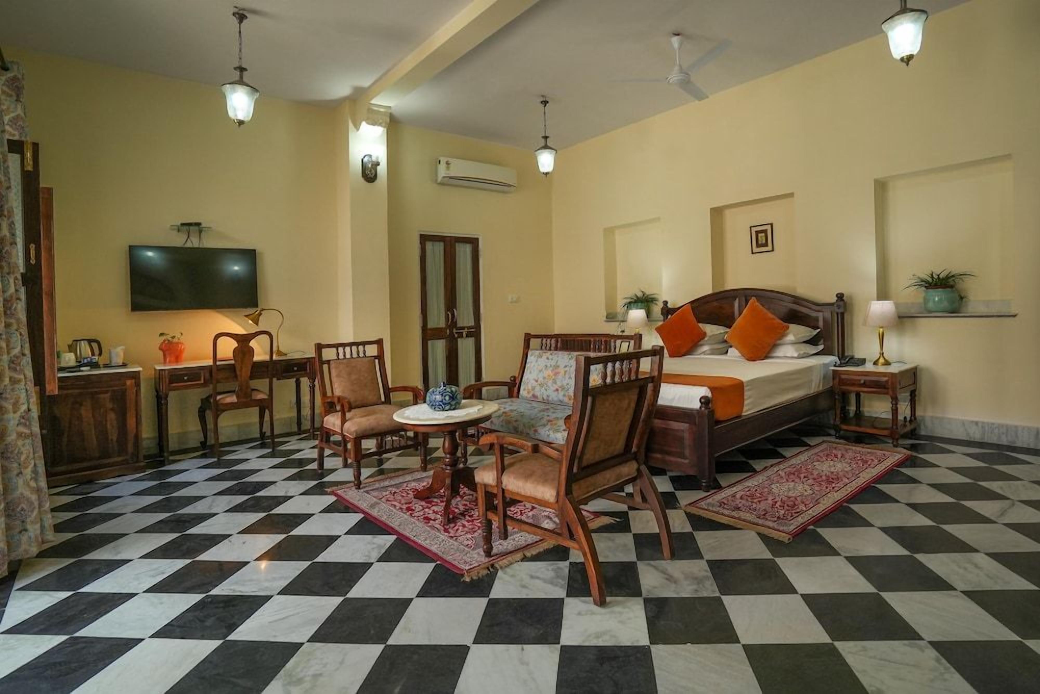Une chambre d’hôtel avec un sol à carreaux, un lit avec des oreillers orange, un bureau avec une chaise, une télévision et un climatiseur fixé au mur.