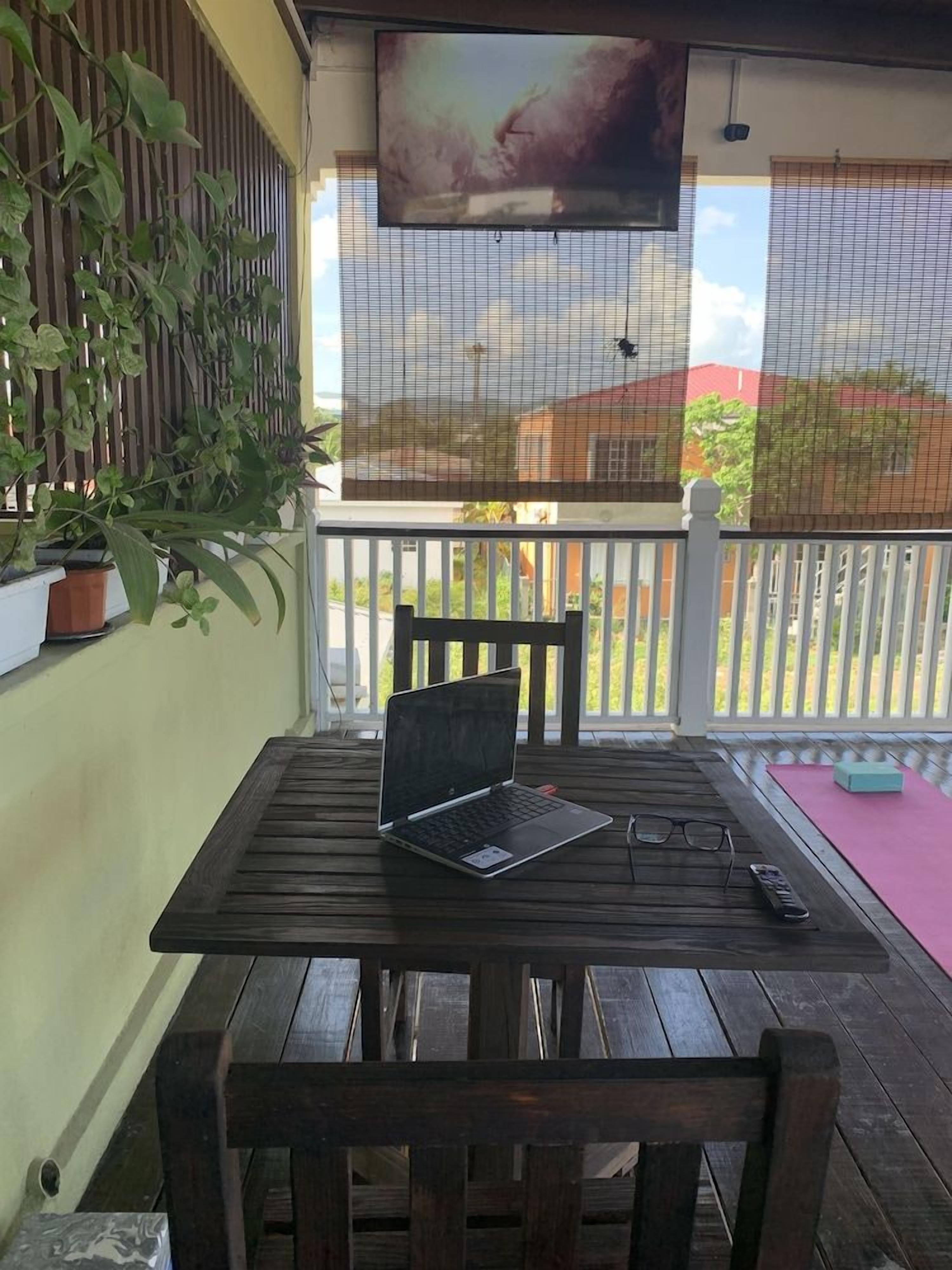 Mesa de madeira com um laptop, óculos e um controle remoto em uma varanda com corrimão branco, plantas em vasos e uma tela de TV.