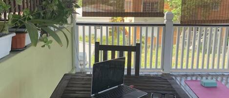 Mesa de madeira com um laptop, óculos e um controle remoto em uma varanda com corrimão branco, plantas em vasos e uma tela de TV.