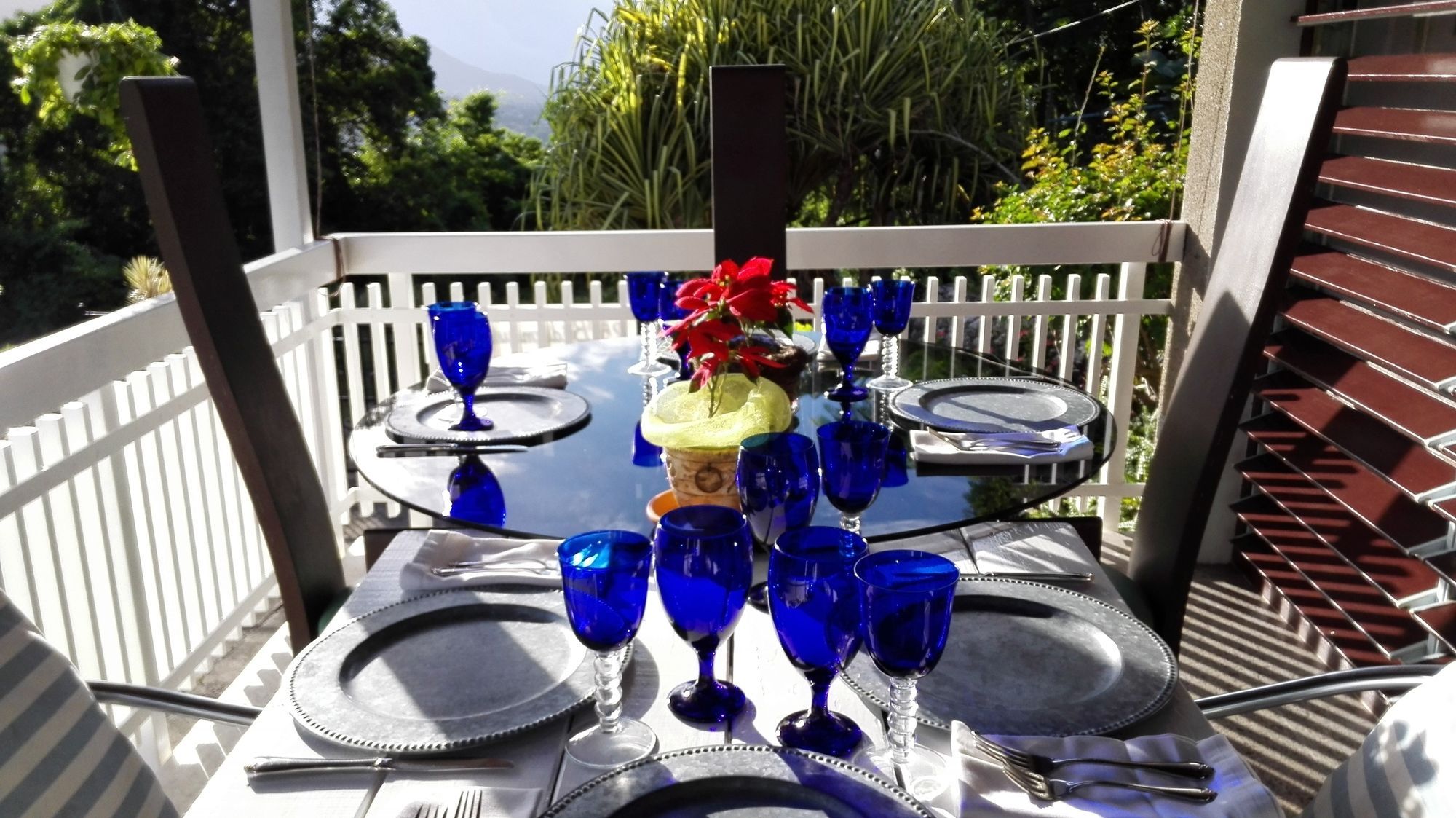 A table set for four with blue glassware, a red flower centerpiece, and a white fence surrounding the area.
