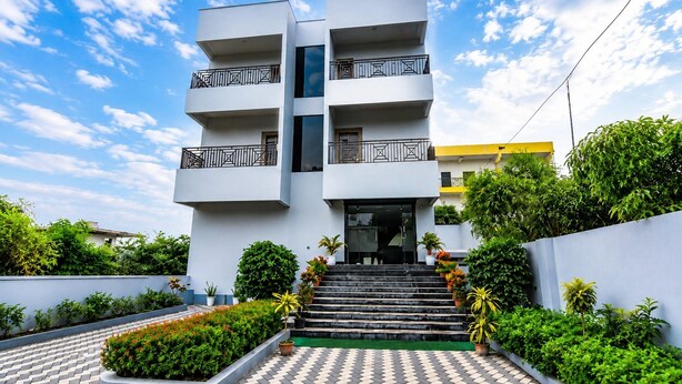 Un edificio de varios pisos con una escalera central que conduce a la entrada, rodeado de follaje y un cielo despejado.