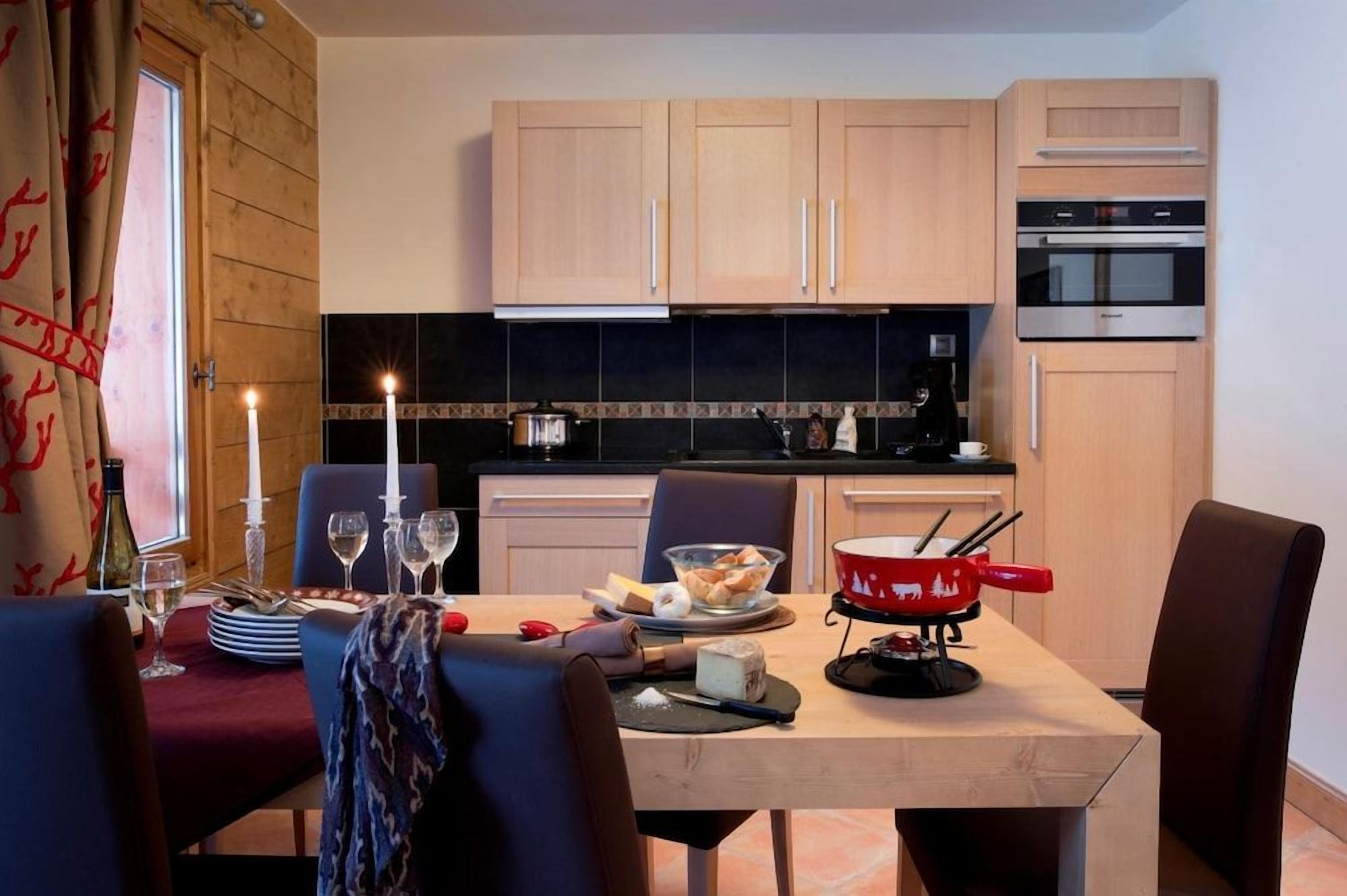 Ein Essbereich mit einem Tisch für vier Personen, einem roten Käsefondue-Set und einer Küche mit Holzschränken und einer schwarzen Spritzschutzwand.