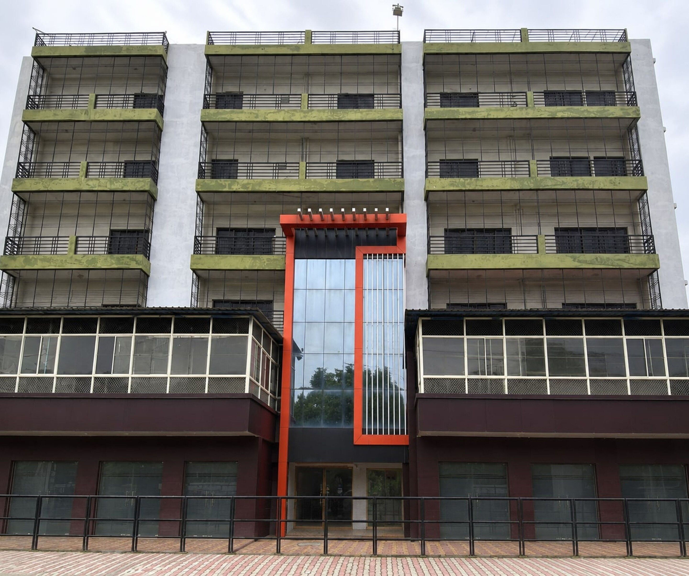 A multi-story building with a central glass entrance, balconies on each floor, and a brick-paved ground.