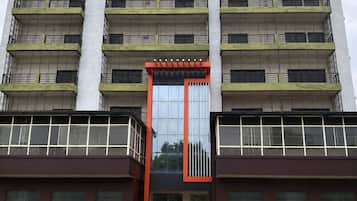 A multi-story building with a central glass entrance, balconies on each floor, and a brick-paved ground.