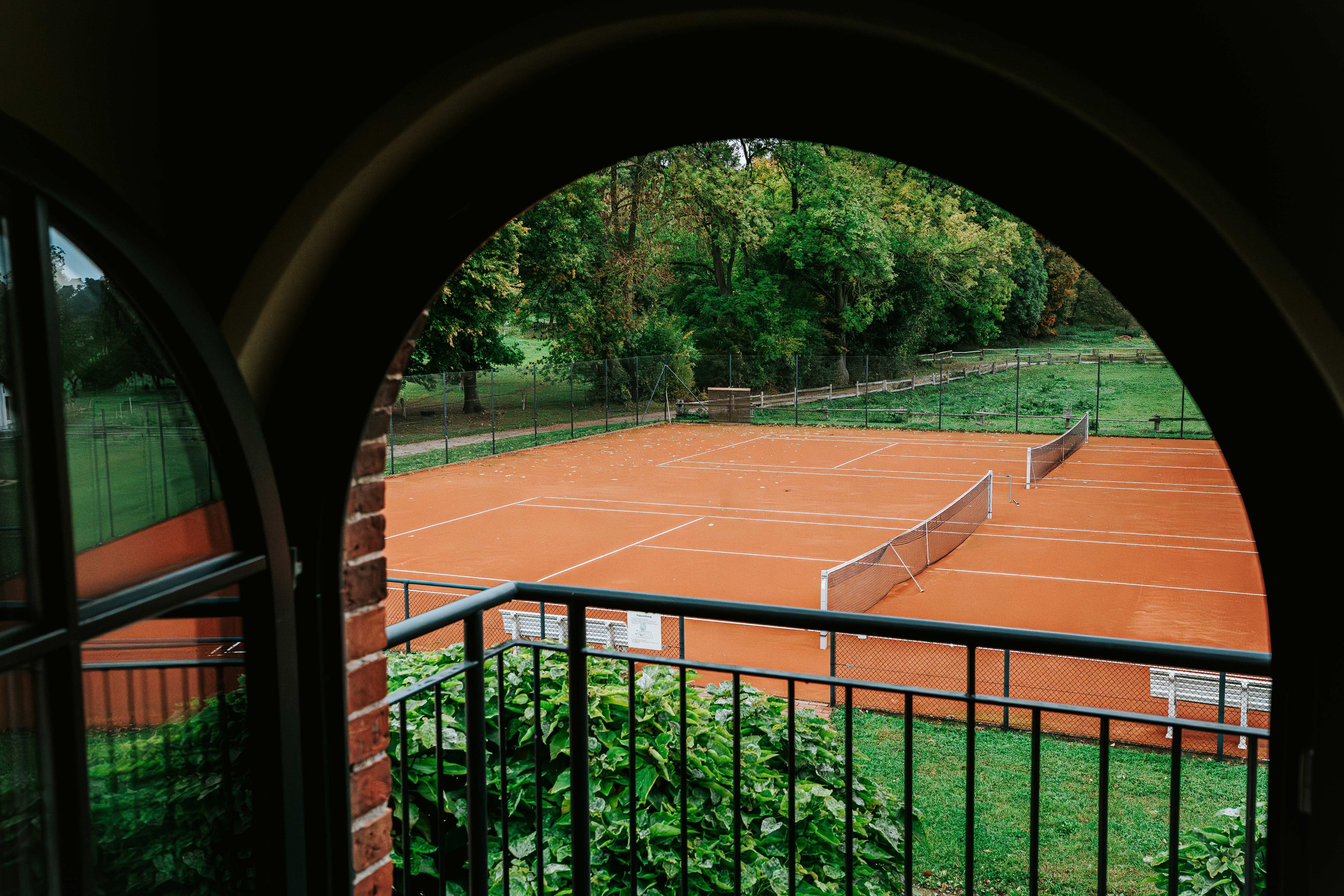 An outdoor tennis court with a brick surface, surrounded by a fence and greenery.