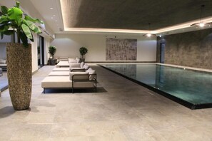 A modern indoor swimming pool area with a lounge, seating, and a large plant.