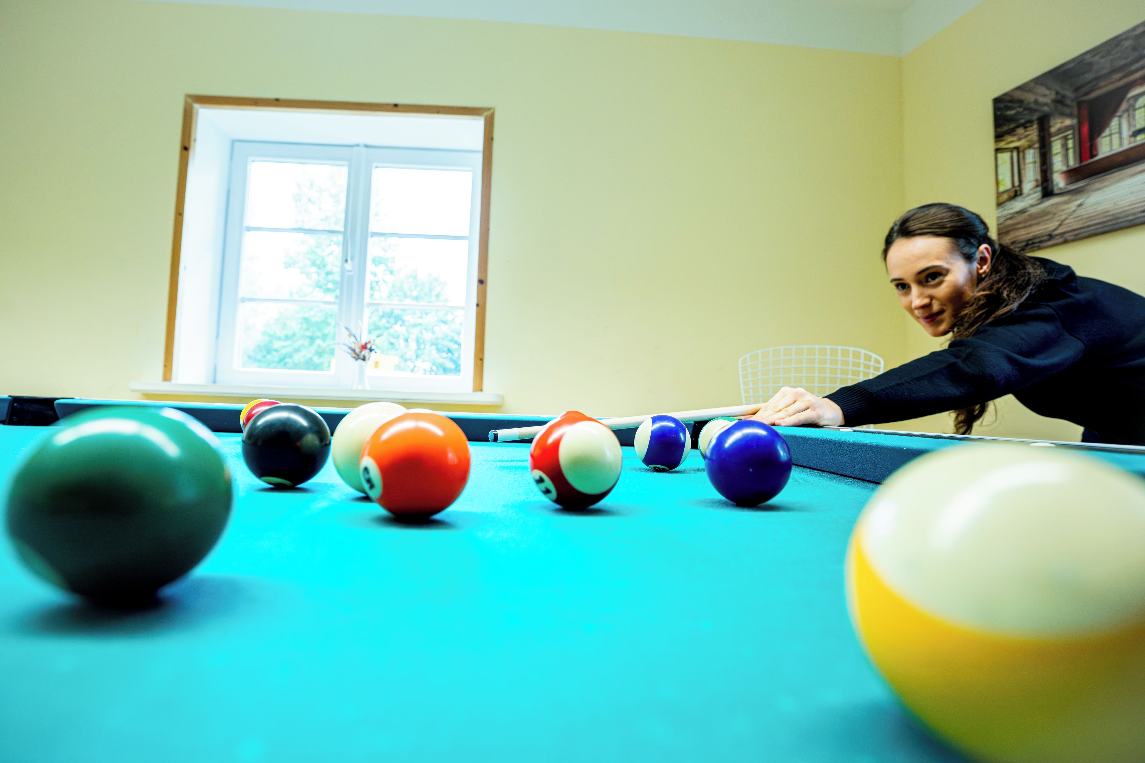 A pool table with balls in a room with a window and a painting on the wall.