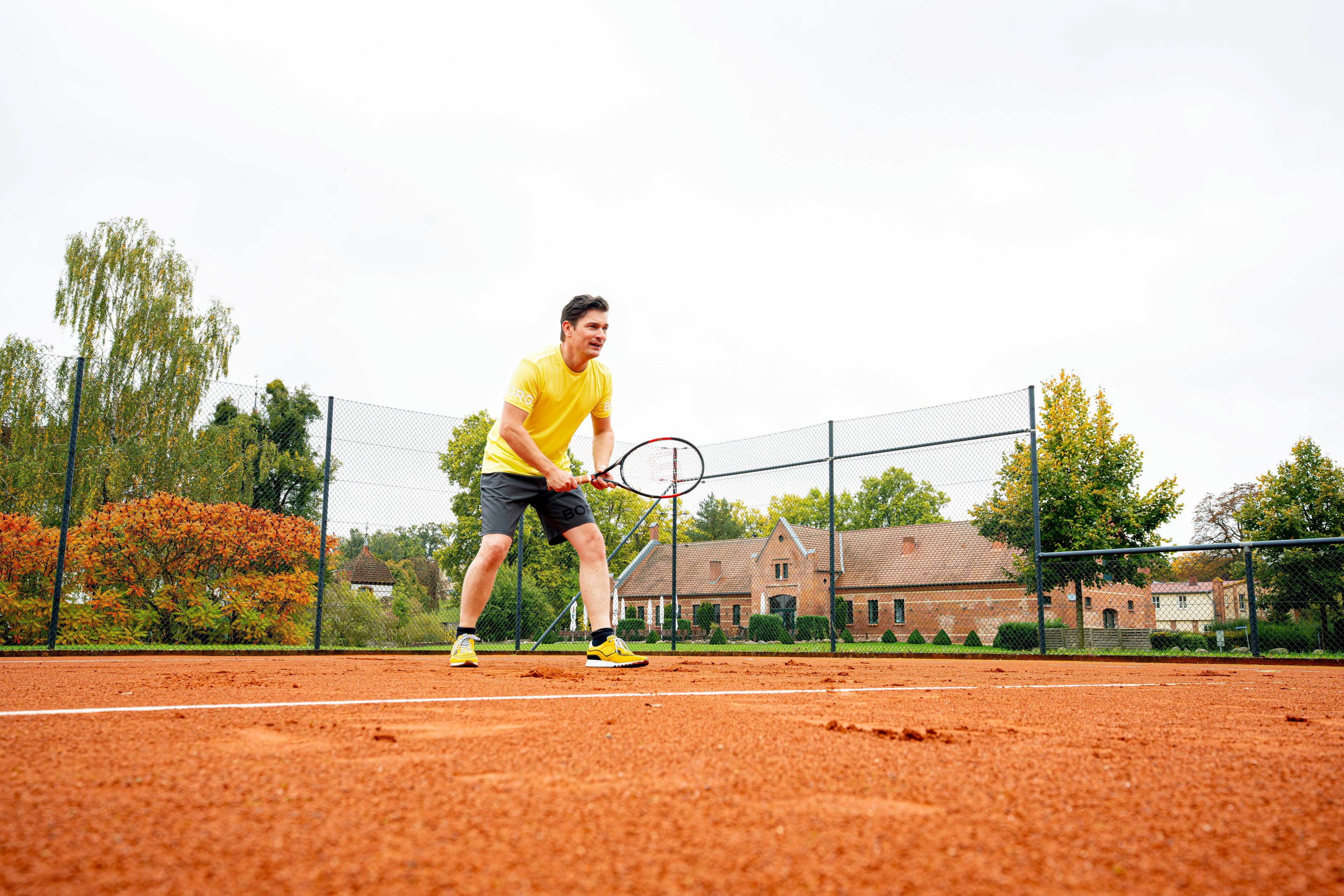 Ein Tennisspieler auf einem Sandplatz mit einem Zaun, Bäumen und Gebäuden im Hintergrund.