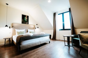 A modern bedroom with a bed, a side table, a chair, a window with curtains, and a painting on the wall.