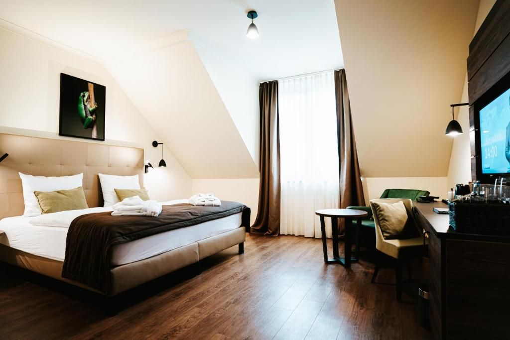 Habitación de hotel con cama, una mesita, una silla, un televisor y una ventana con cortinas.