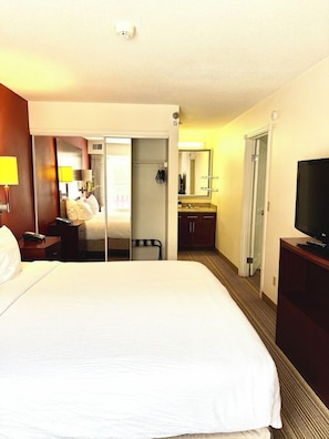 Habitación de hotel con una cama grande, televisión, escritorio con lámpara, espejo, baño con lavamanos y ducha.
