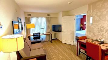 Quarto de hotel com sofá, mesa de jantar, televisão e cama visível por uma porta aberta.