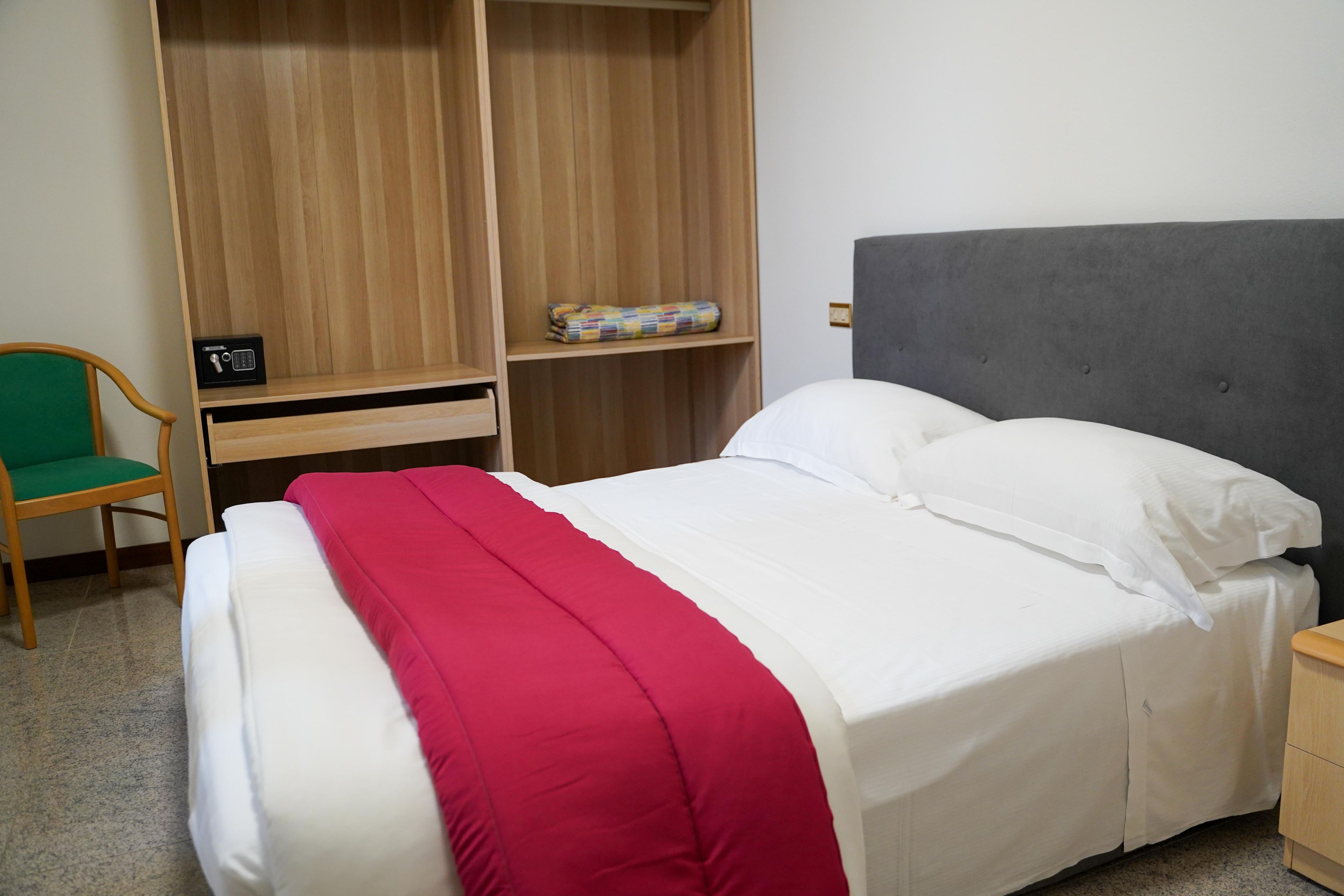 A neatly made bed with white and red bedding, a grey upholstered headboard, a wooden wardrobe, a small desk with a radio, and a green chair.