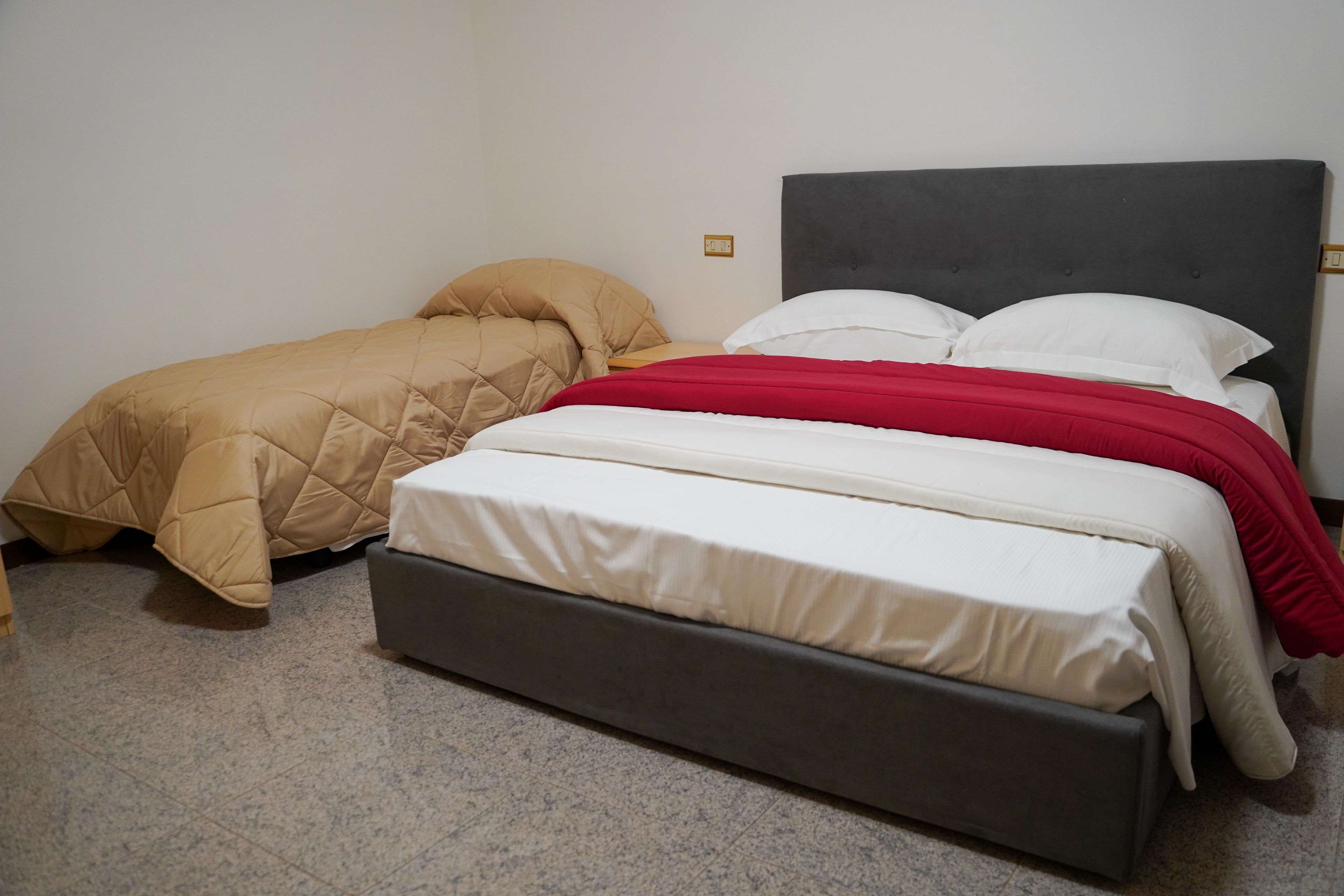 Een tweepersoonsbed met een grijs hoofdeinde en een rode deken, een eenpersoonsbed met een beige sprei, en een vloer met grijze vloerbedekking.