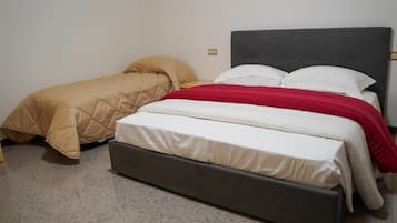 Een tweepersoonsbed met een grijs hoofdeinde en een rode deken, een eenpersoonsbed met een beige sprei, en een vloer met grijze vloerbedekking.