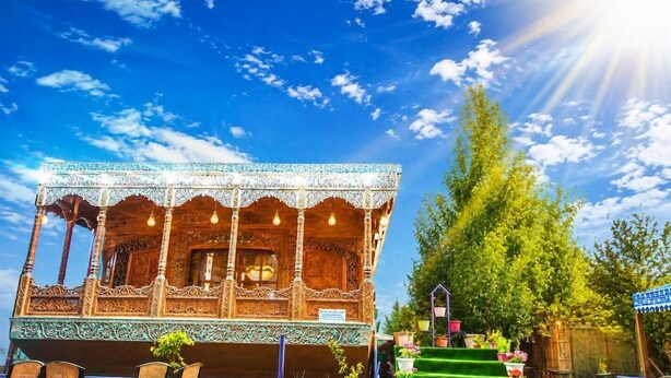 A traditional wooden building with intricate carvings, a balcony with wicker furniture, and a staircase leading to a green lawn area. - De - Laila Group Of House Boats (Srinagar)