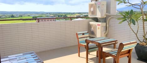 An outdoor balcony with a view of a green landscape, a table and chairs, and two air conditioning units.